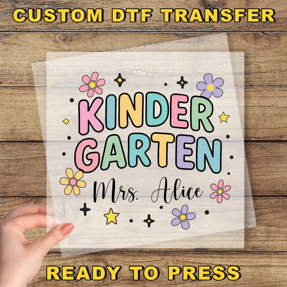 A hand is holding a clear sheet with a colorful design featuring the text "KINDER GARTEN" surrounded by flowers and stars.
