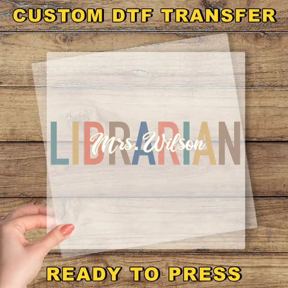The image is a promotional image for a custom DTF (Digital Transfer Factory) transfer service. It features a hand holding a clear sheet with the word "LIBRARIAN" written on it, placed on a wooden background. The text "CUSTOM DTF TRANSFER" is displayed at the top of the image, and "READY TO PRESS" is written at the bottom.