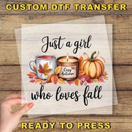 The image is a hand holding a transparent sheet with a design featuring a candle, a mug with a leaf, and a pumpkin surrounded by autumn leaves. The text on the sheet reads "Just a girl who loves fall" and "Ready to press".