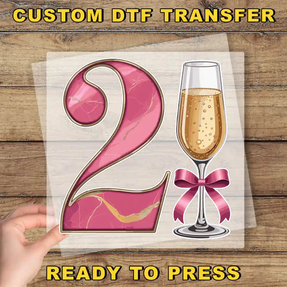 a hand holding a transparent sheet with a pink number "2" and a glass of champagne with a bow on it.