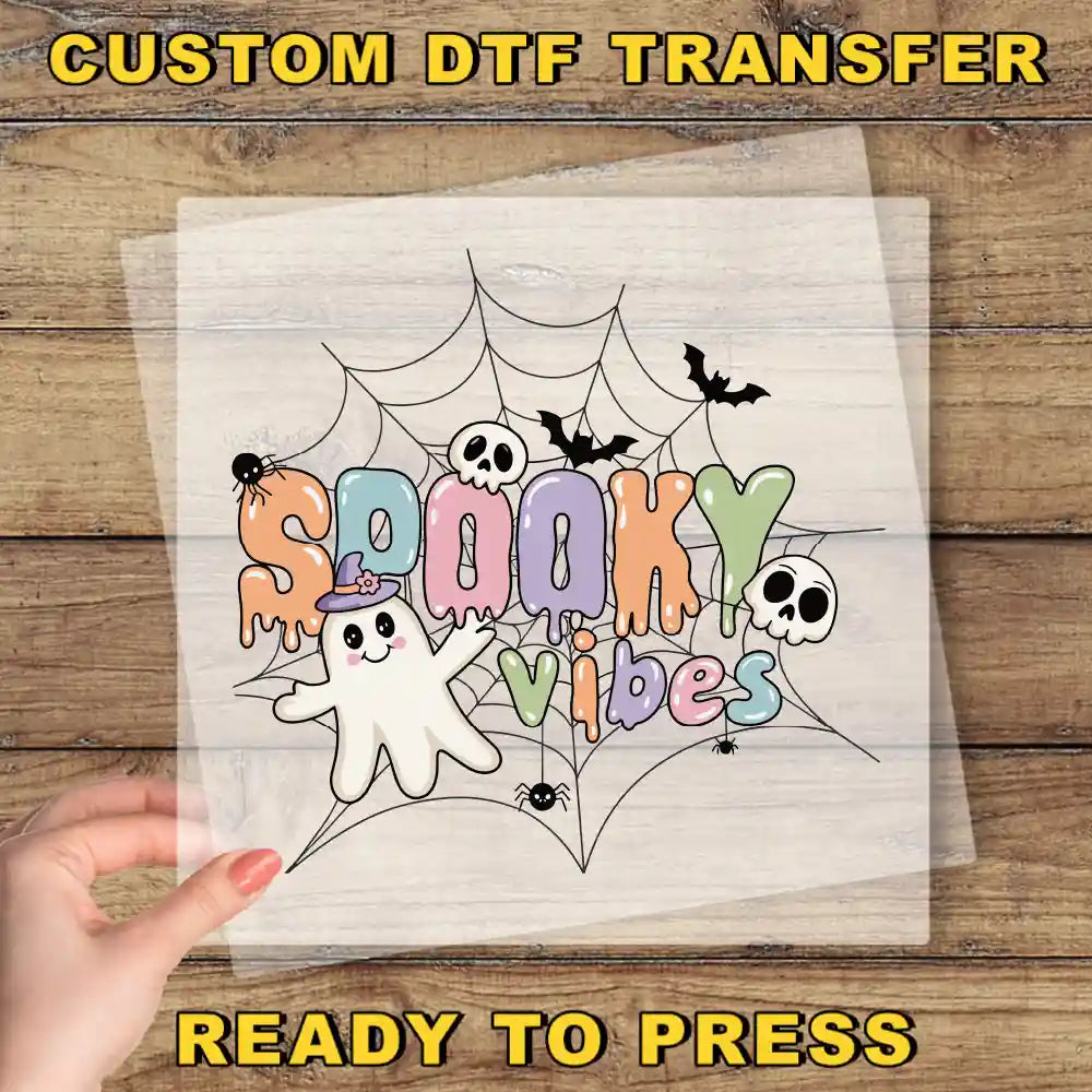 A hand is holding a sheet of paper with a Halloween-themed design featuring a ghost, spider, bats, and the text "SPOOKY VIBES" in a colorful, spooky font.