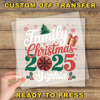 A hand is holding a transparent sheet with a festive Christmas design featuring text and decorative elements such as a tree, snowflakes, and a gift box.