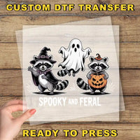 A hand is holding a clear sheet with a Halloween-themed illustration of a raccoon dressed as a witch and a ghost, along with a pumpkin.