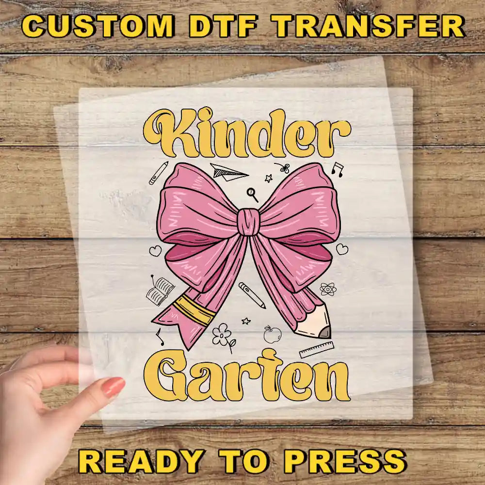The image is a hand holding a transparent sheet with a pink bow and various school supplies, including a pencil and ruler, along with the text "Kinder Garten" and "READY TO PRESS".