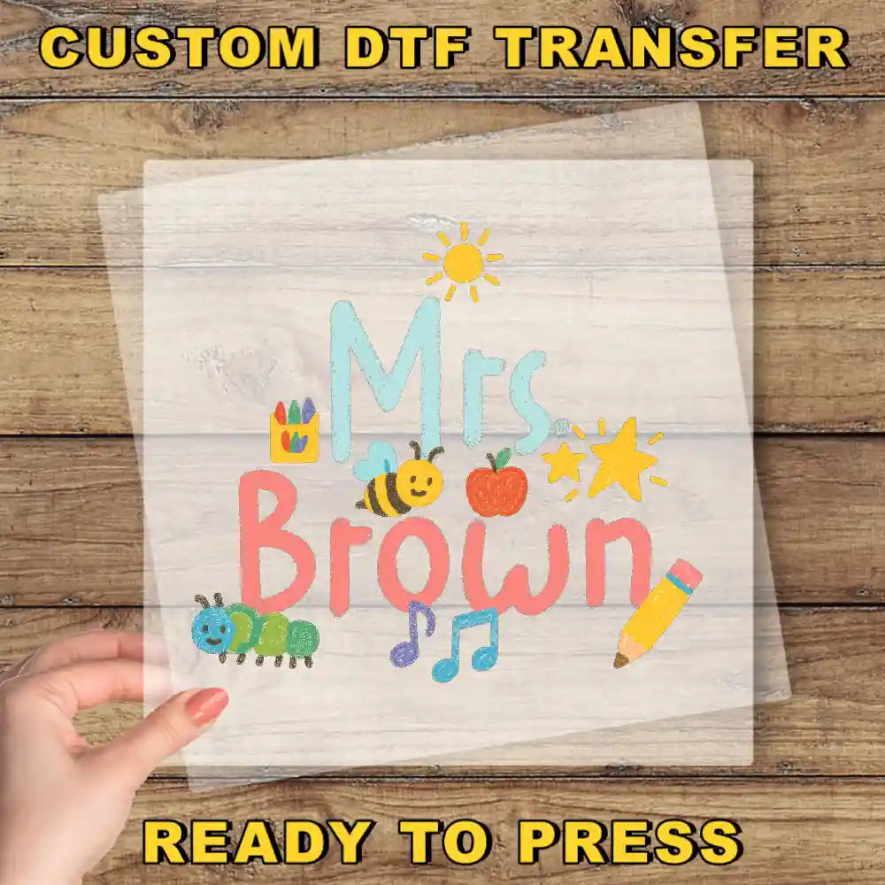 The image is a hand holding a piece of paper with a colorful design featuring the text "Mrs Brown" surrounded by various school-related objects such as a pencil, apple, and bee.