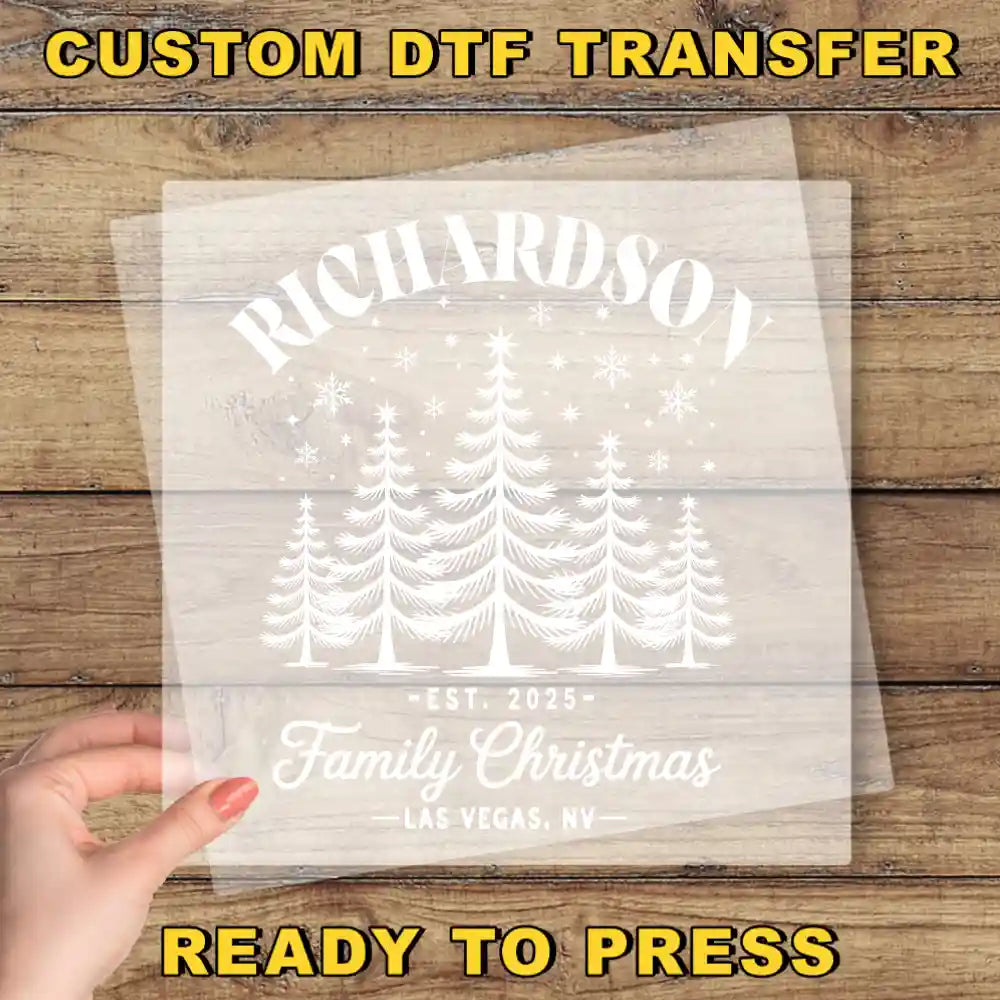 The image is a hand holding a transparent sheet with a white design featuring a Christmas tree and snowflakes. The text on the sheet reads "RICHARDSON Family Christmas Las Vegas, Nevada" and "CUSTOM DTF TRANSFER".