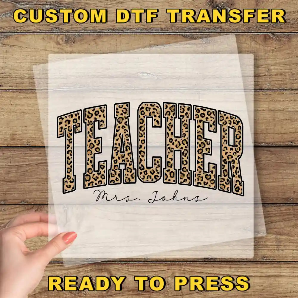 A hand holding a clear sheet with the word "TEACHER" printed on it, along with the name "Mrs. Johns" written below it.