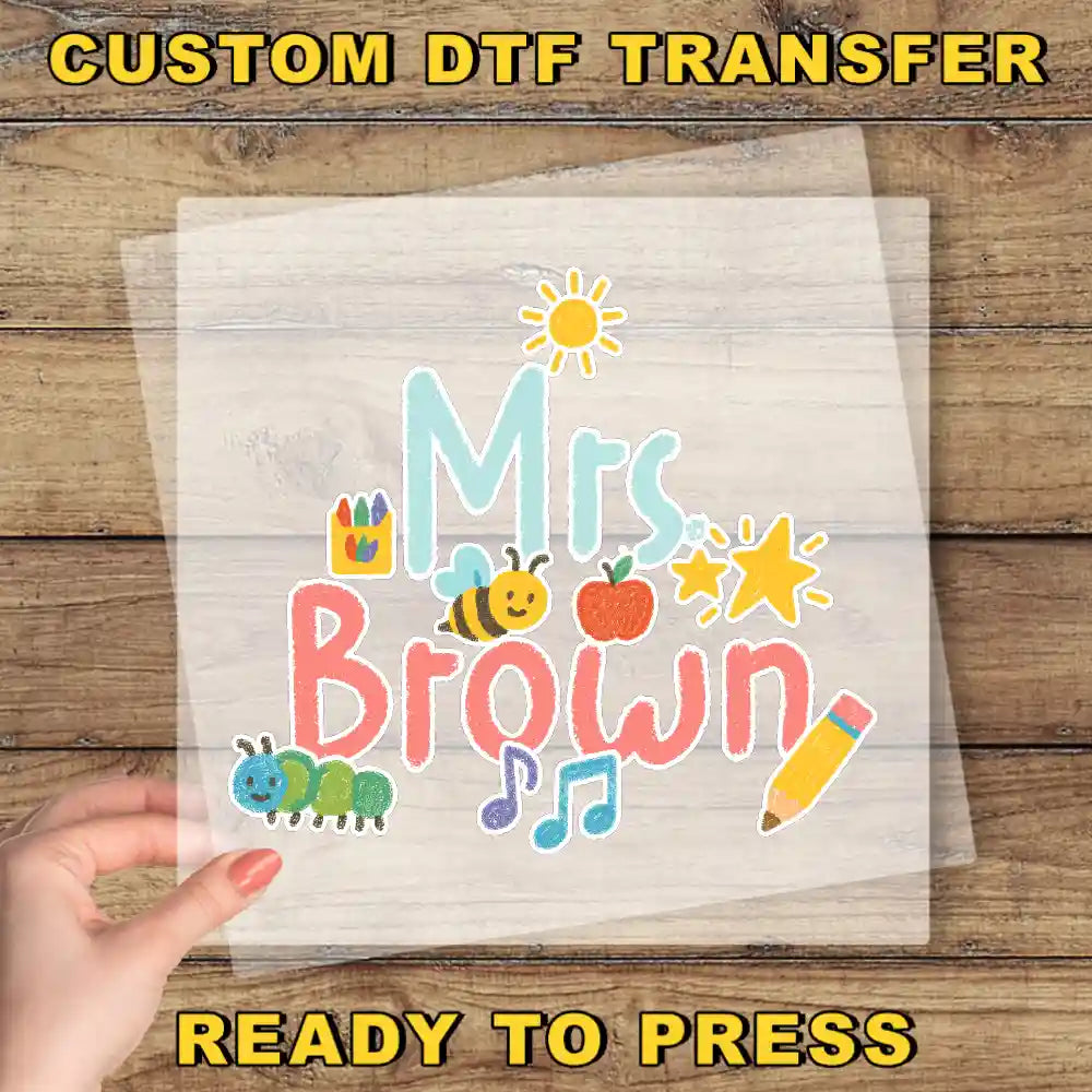 A hand is holding a transparent sheet with a colorful design featuring the text "Mrs. Brown" and various cartoon-like illustrations such as a bee, apple, and pencil.