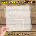 A hand holding a transparent sheet with the text "Custom Dtf Transfer" and "image" written on it, against a wooden background.