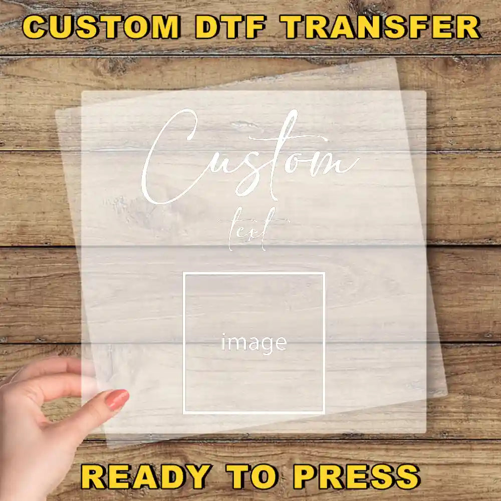 A hand holding a transparent sheet with the text "Custom Dtf Transfer" and "image" written on it, against a wooden background.