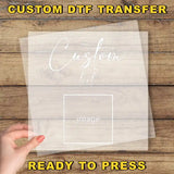 A hand holding a transparent sheet with the text "Custom Dtf Transfer" and "image" written on it, against a wooden background.