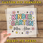 A hand is holding a clear sheet with a colorful design featuring the text "KINDER GARTEN" surrounded by flowers and stars.