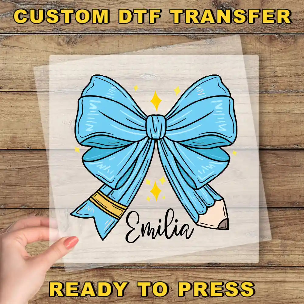 a hand holding a clear plastic sheet with a blue bow and a pencil, with the name "Emilia" written on it.