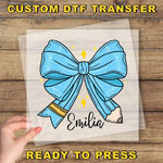 a hand holding a clear plastic sheet with a blue bow and a pencil, with the name "Emilia" written on it.