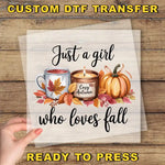The image is a hand holding a transparent sheet with a design featuring a candle, a mug with a leaf, and a pumpkin surrounded by autumn leaves. The text on the sheet reads "Just a girl who loves fall" and "Ready to press".
