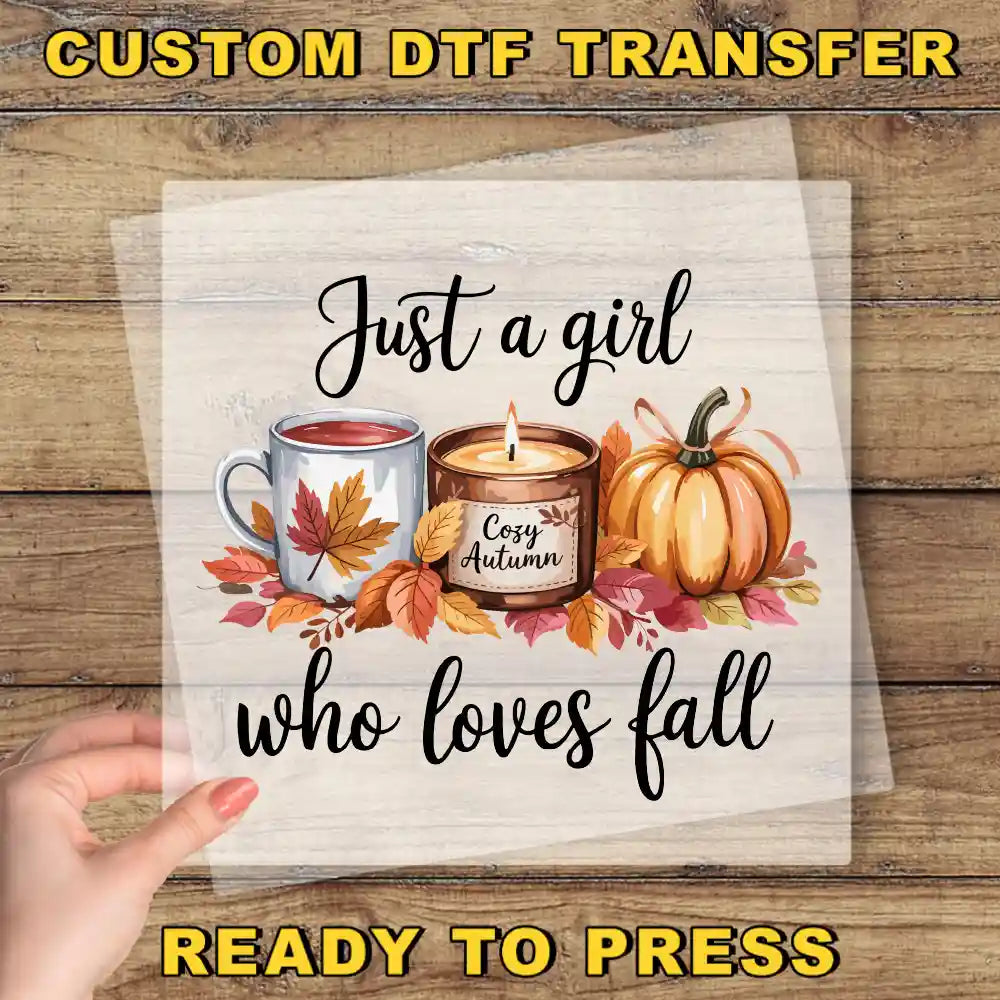 The image is a hand holding a transparent sheet with a design featuring a candle, a mug with a leaf, and a pumpkin surrounded by autumn leaves. The text on the sheet reads "Just a girl who loves fall" and "Ready to press".