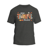 A gray t-shirt with a graphic design featuring the text "One Thankful Teacher" surrounded by autumn-themed elements such as leaves, pumpkins, and flowers.