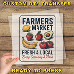 The image is a poster for a farmers market, featuring various fruits and vegetables such as bananas, apples, and avocados. The poster is displayed on a wooden surface, and there is a hand holding it up, suggesting that it is ready to be printed or displayed.