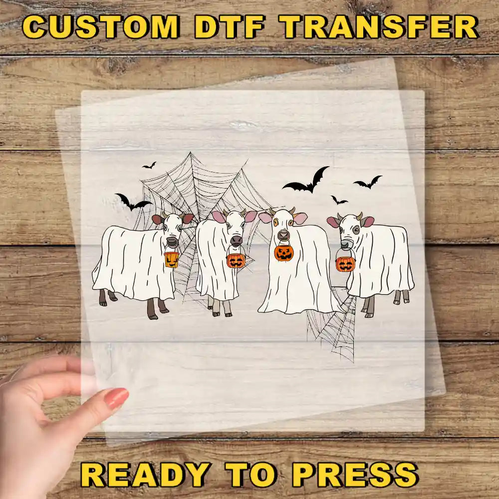 a hand holding a transparent sheet with a Halloween-themed illustration of cows dressed in ghost costumes, with bats flying around them.