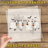 a hand holding a transparent sheet with a Halloween-themed illustration of cows dressed in ghost costumes, with bats flying around them.
