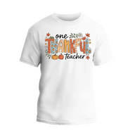 A white t-shirt with a graphic design featuring the text "One Thankful Teacher" surrounded by autumn-themed elements such as leaves, pumpkins, and flowers.