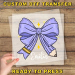 a hand holding a transparent sheet with a purple bow and a pencil, with the name "Emilia" written on it.