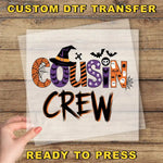 a hand holding a piece of paper with the text "Cousin Crew" written on it, along with various Halloween-themed decorations such as bats, skulls, and a spider web.