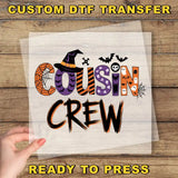 a hand holding a piece of paper with the text "Cousin Crew" written on it, along with various Halloween-themed decorations such as bats, skulls, and a spider web.