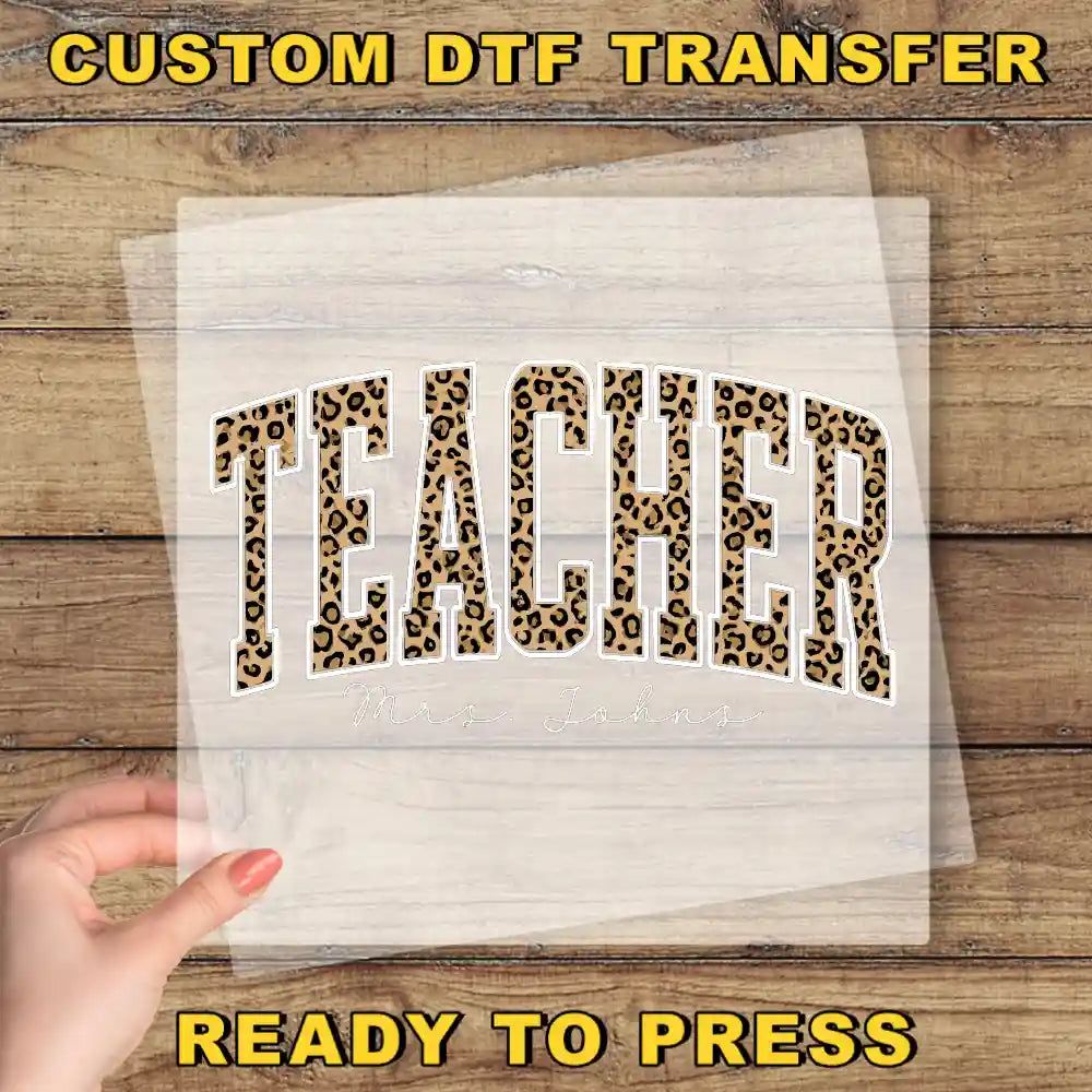 a hand holding a transparent sheet with the word "TEACHER" printed on it, set against a wooden background.