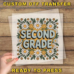 a hand holding a piece of paper with a floral design and the text "Second Grade" printed on it.
