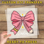 a hand holding a transparent sheet with a pink bow and a pencil, with the name "Emilia" written on it.