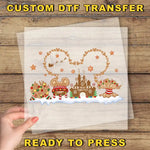 A hand is holding a clear sheet with a festive winter scene featuring various Disney-themed elements such as Mickey Mouse, a gingerbread house, and a snowman.