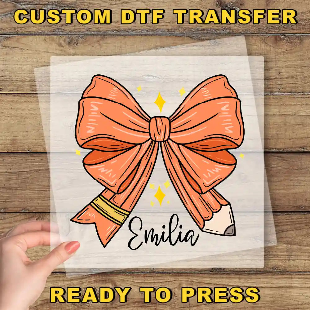a hand holding a clear sheet with a large orange bow and a pencil, with the name "Emilia" written below it.