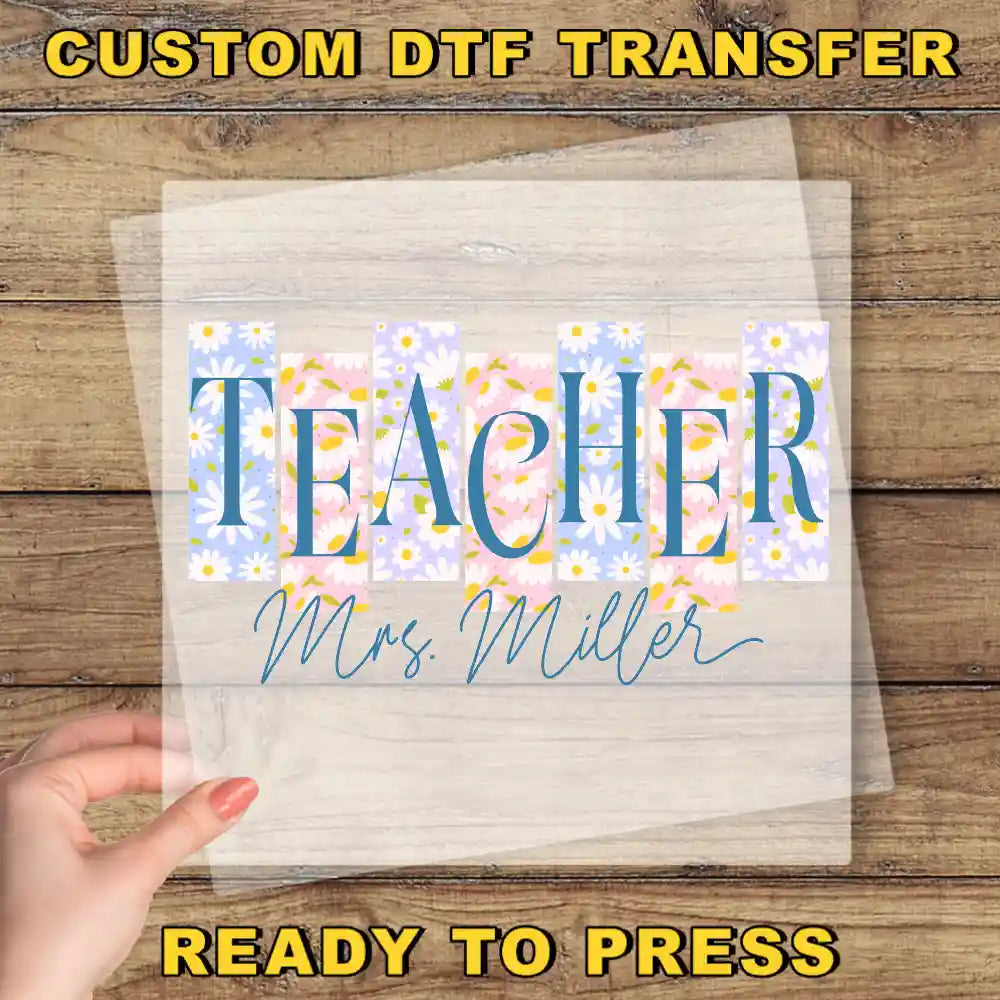 The image is a promotional image for a custom DTF transfer service, featuring a hand holding a clear sheet with the text "Teacher Mrs. Miller" printed on it. The text is surrounded by a floral pattern, and the background is a wooden surface.