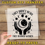 The image is a hand holding a transparent sheet with a graphic design featuring a crescent moon, stars, and the text "They didn't burn witches, they burned women".