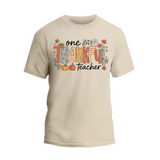 A beige t-shirt with a graphic design featuring the text "One Thankful Teacher" surrounded by autumn-themed elements such as leaves, pumpkins, and flowers.