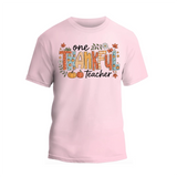 A pink t-shirt with a graphic design featuring the text "One Thankful Teacher" surrounded by autumn-themed elements such as leaves, pumpkins, and flowers.