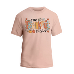 A light pink t-shirt with a graphic design featuring the text "One Thankful Teacher" surrounded by autumn-themed elements such as leaves, pumpkins, and flowers.