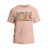 A light pink t-shirt with a graphic design featuring the text "One Thankful Teacher" surrounded by autumn-themed elements such as leaves, pumpkins, and flowers.