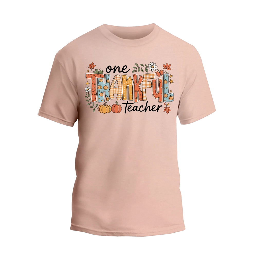 A light pink t-shirt with a graphic design featuring the text "One Thankful Teacher" surrounded by autumn-themed elements such as leaves, pumpkins, and flowers.