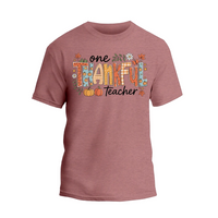 A pink t-shirt with a graphic design featuring the text "One Thankful Teacher" surrounded by autumn-themed elements such as leaves, pumpkins, and flowers.