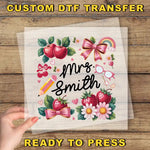 The image is a hand holding a transparent sheet with a colorful illustration of various objects, including apples, flowers, and a pencil, along with the text "Mrs. Smith" and "Ready to Press".