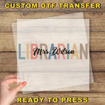 a hand holding a clear sheet with the word "LIBRARIAN" written on it, placed on a wooden surface.