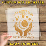a hand holding a transparent sheet with a graphic design on it. The design includes a crescent moon, stars, and the text "They didn't burn witches, they burned women".