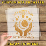 a hand holding a transparent sheet with a graphic design on it. The design includes a crescent moon, stars, and the text "They didn't burn witches, they burned women".