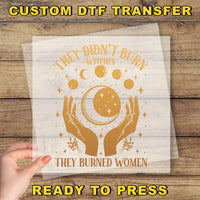 a hand holding a transparent sheet with a graphic design on it. The design includes a crescent moon, stars, and the text "They didn't burn witches, they burned women".