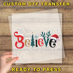 A hand holding a clear sheet with the word "Believe" written on it, surrounded by snowflakes and Christmas-themed decorations.