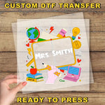 A hand holding a transparent sheet with a colorful illustration of a classroom scene, including a globe, books, a laptop, and various school supplies.