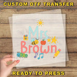 The image is a hand holding a piece of paper with a colorful design featuring the text "Mrs Brown" surrounded by various school-related objects such as a pencil, apple, and bee.