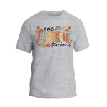 A gray t-shirt with a graphic design featuring the text "One Thankful Teacher" surrounded by autumn-themed elements such as leaves, pumpkins, and flowers.